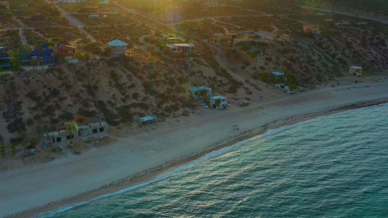Aerial frame over El Sargento La Ventana, Mexico, showing textured blue water in the lower right, a diagonal beach, hillside scrub, several buildings, and no visible people.