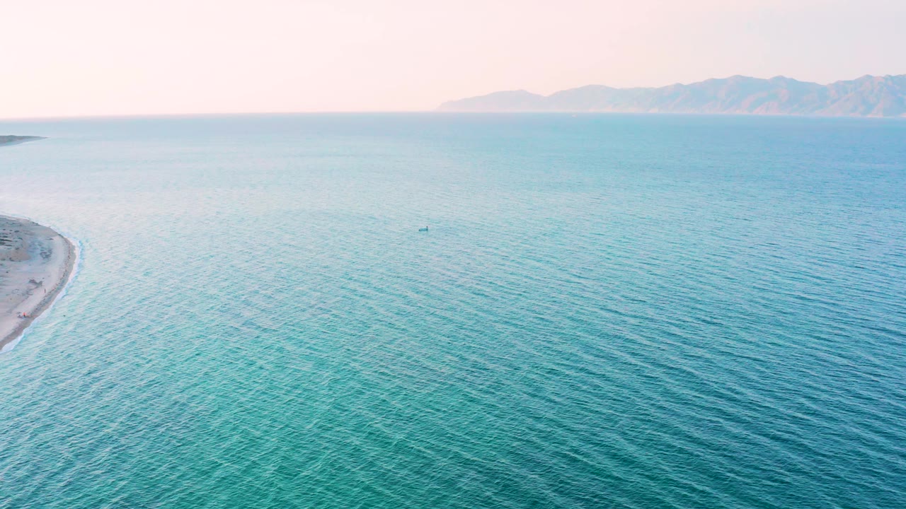 High aerial view in El Sargento La Ventana, Mexico with blue-green water across the foreground, a small dark boat near the middle distance, hazy mountains beyond, and no visible people.