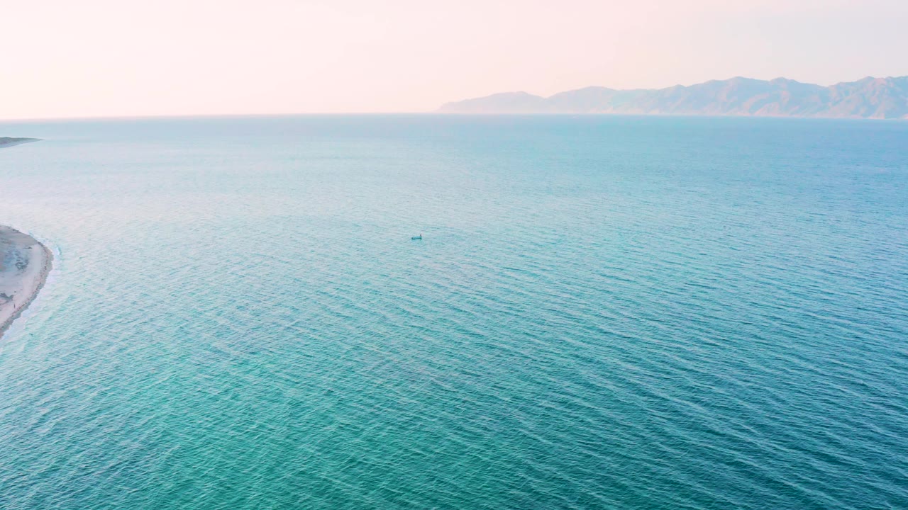 Wide drone frame from El Sargento La Ventana, Mexico with calm blue water, sandy beach visible along the left side, a small boat offshore, soft horizon haze, and no visible people.