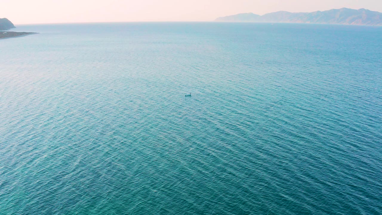 Opening frame over turquoise water near El Sargento La Ventana, Mexico, with faint mountains on the horizon, a small dark boat or floating object offshore, and no visible people.