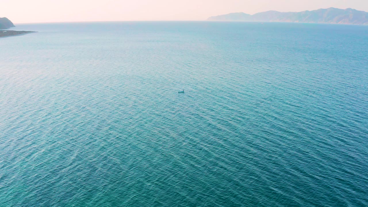 Aerial frame of rippled blue water in El Sargento La Ventana, Mexico, with a small offshore shape near center and hazy mountain silhouettes across the background, no visible people.