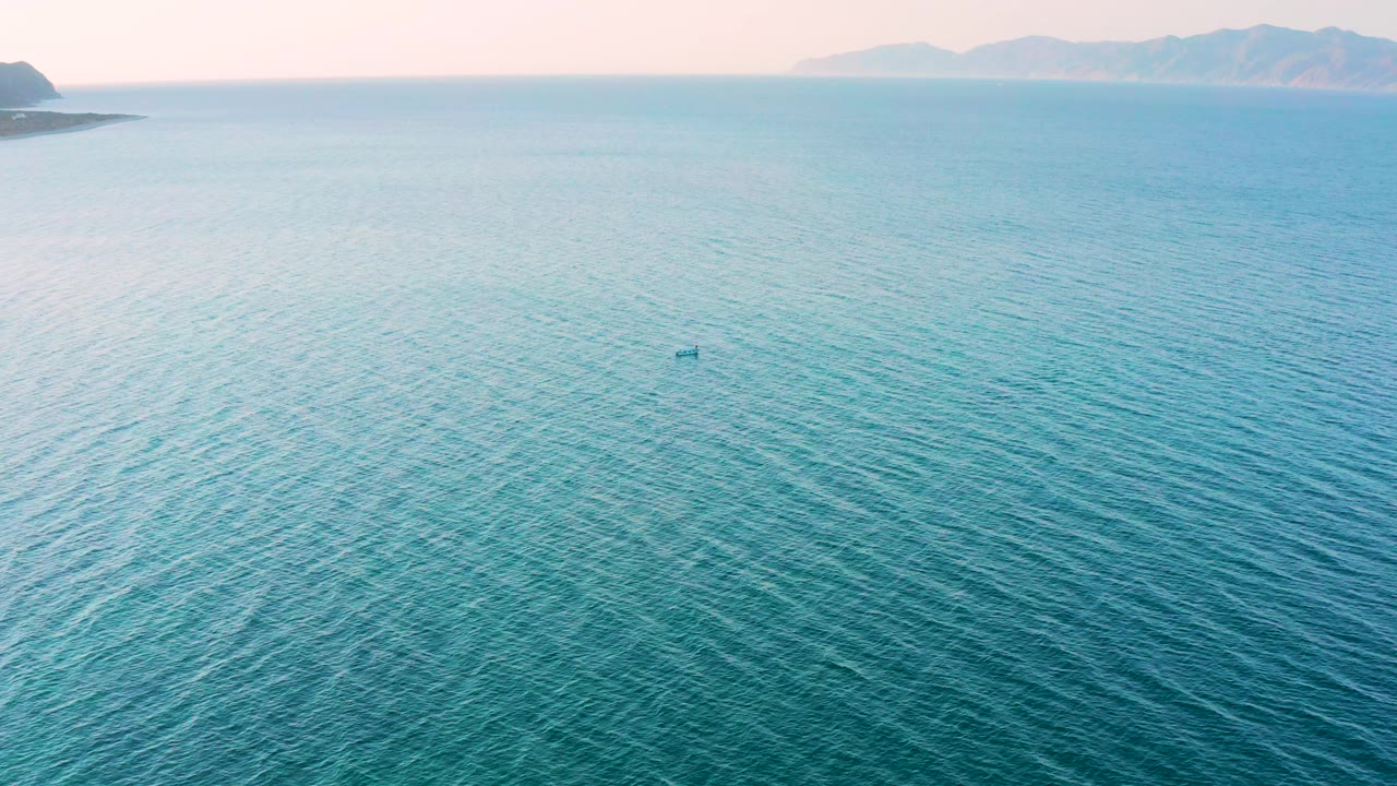 Wide blue water frame from El Sargento La Ventana, Mexico, with the small dark offshore object slightly right of center, distant land and mountains along the top edge, and no visible people.