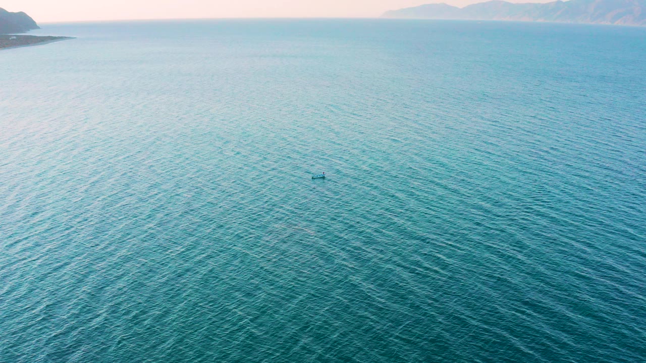 Wide aerial frame over turquoise water in El Sargento La Ventana, Mexico, with a small kayak near center, distant land along the top edge, and no visible people.