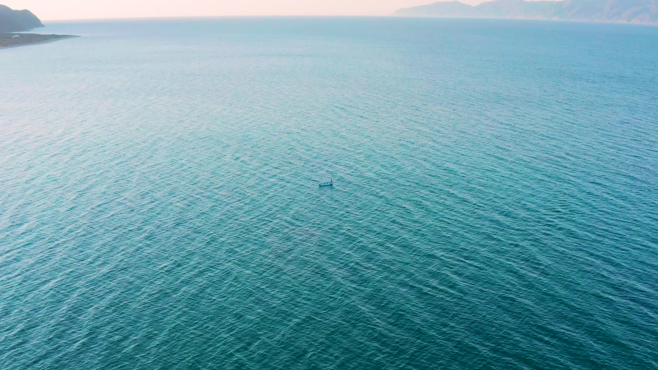 High aerial frame over blue-green water in El Sargento La Ventana, Mexico, showing a tiny kayak near mid-frame, low islands at the horizon, and no visible people.