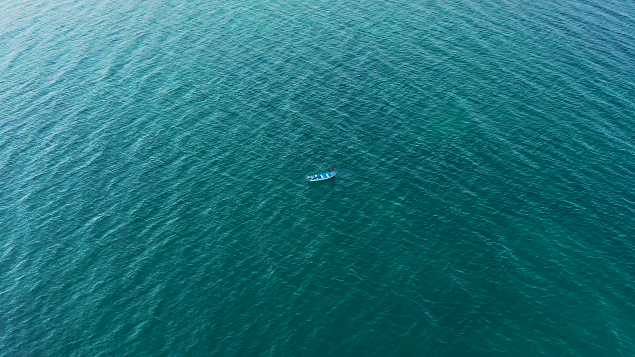 The small fishing boat is still near the top of the frame against darker turquoise sea in El Sargento La Ventana, Mexico, with no visible people.
