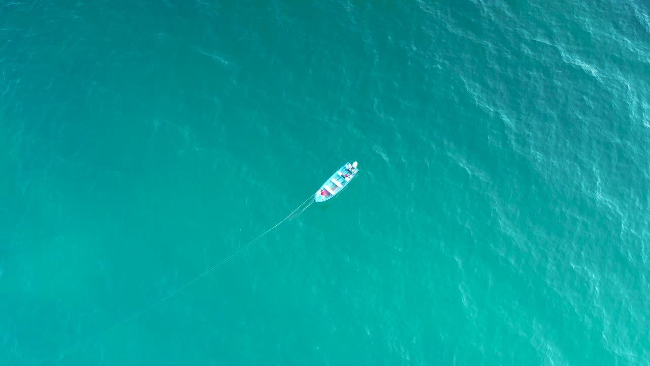 A small fishing boat sits near the upper part of a vertical aerial view, crossing blue-green ocean water with pale wind texture and no visible people.