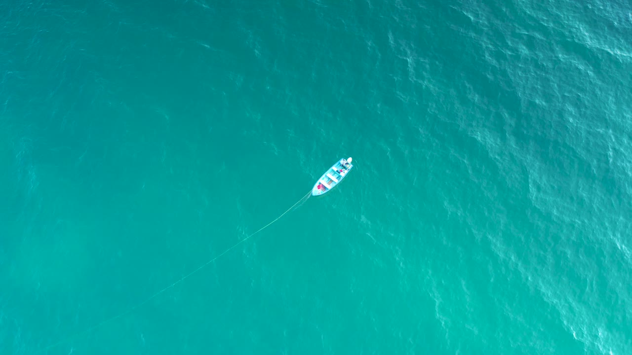 A high overhead view shows the small boat farther across the blue ocean surface, with a thin wake line bending through the water and no visible people.