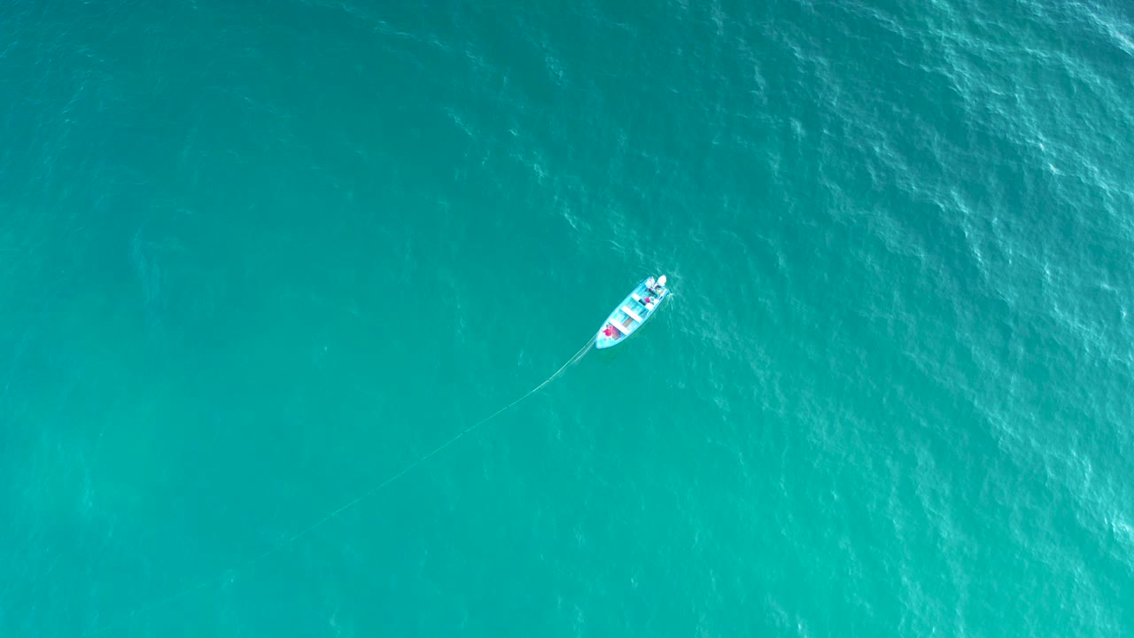 Overhead frame of a small white sailboat near the upper left on turquoise water in El Sargento La Ventana, Mexico, with faint wake and no visible people.