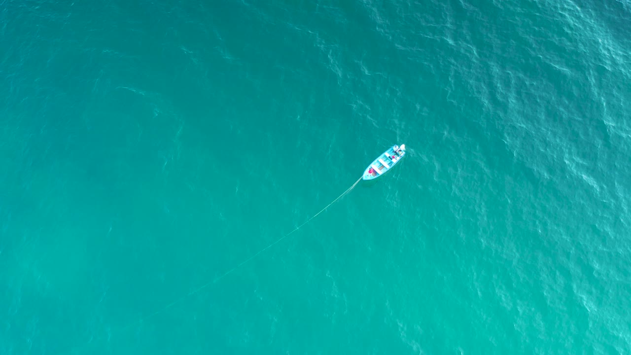 A high aerial view shows the white boat near the upper right of teal water, with darker rippled patches below and no visible people.