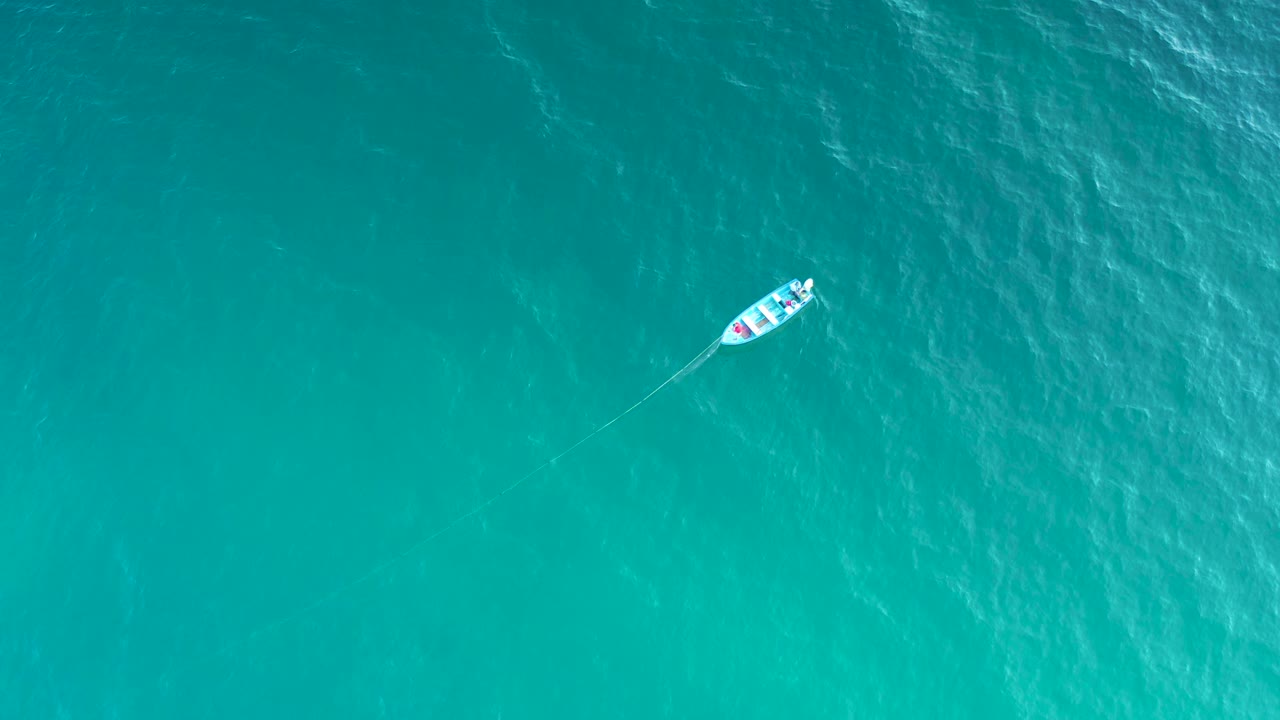 A tiny boat remains near the top of the overhead frame over teal ocean in El Sargento La Ventana, Mexico, with soft surface texture and no visible people.
