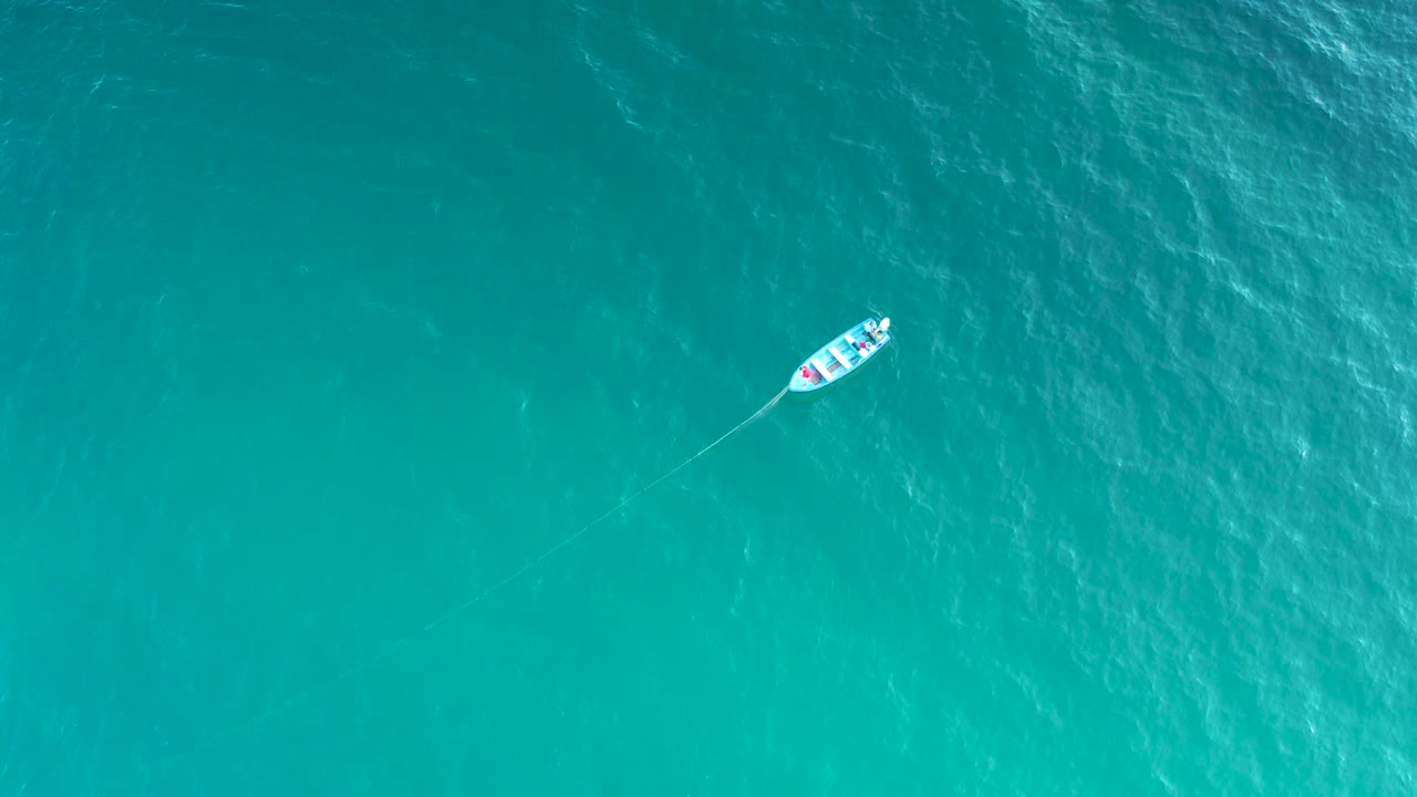 Overhead aerial frame showing the small boat closer to the upper right over bright turquoise sea in El Sargento La Ventana, Mexico, with no shoreline and no visible people.
