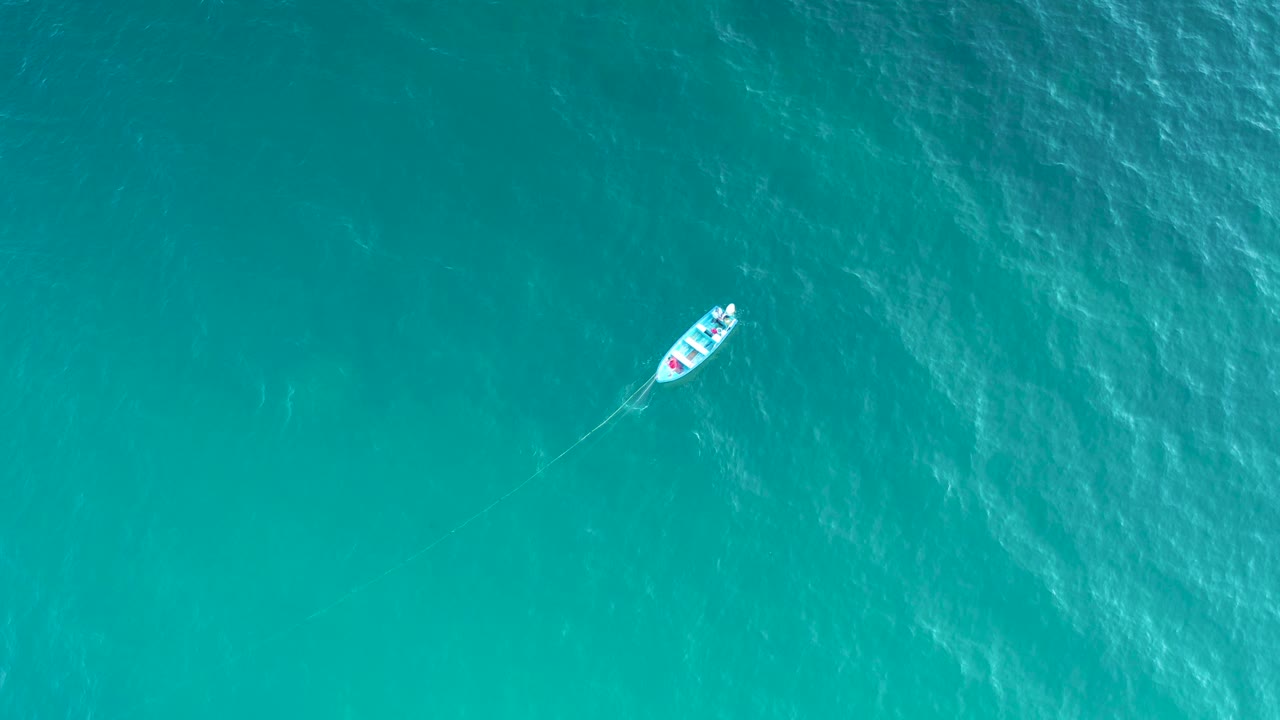 Overhead turquoise sea in El Sargento La Ventana, Mexico, with the small white boat near the upper right and a thin trailing line or wake, no visible people.