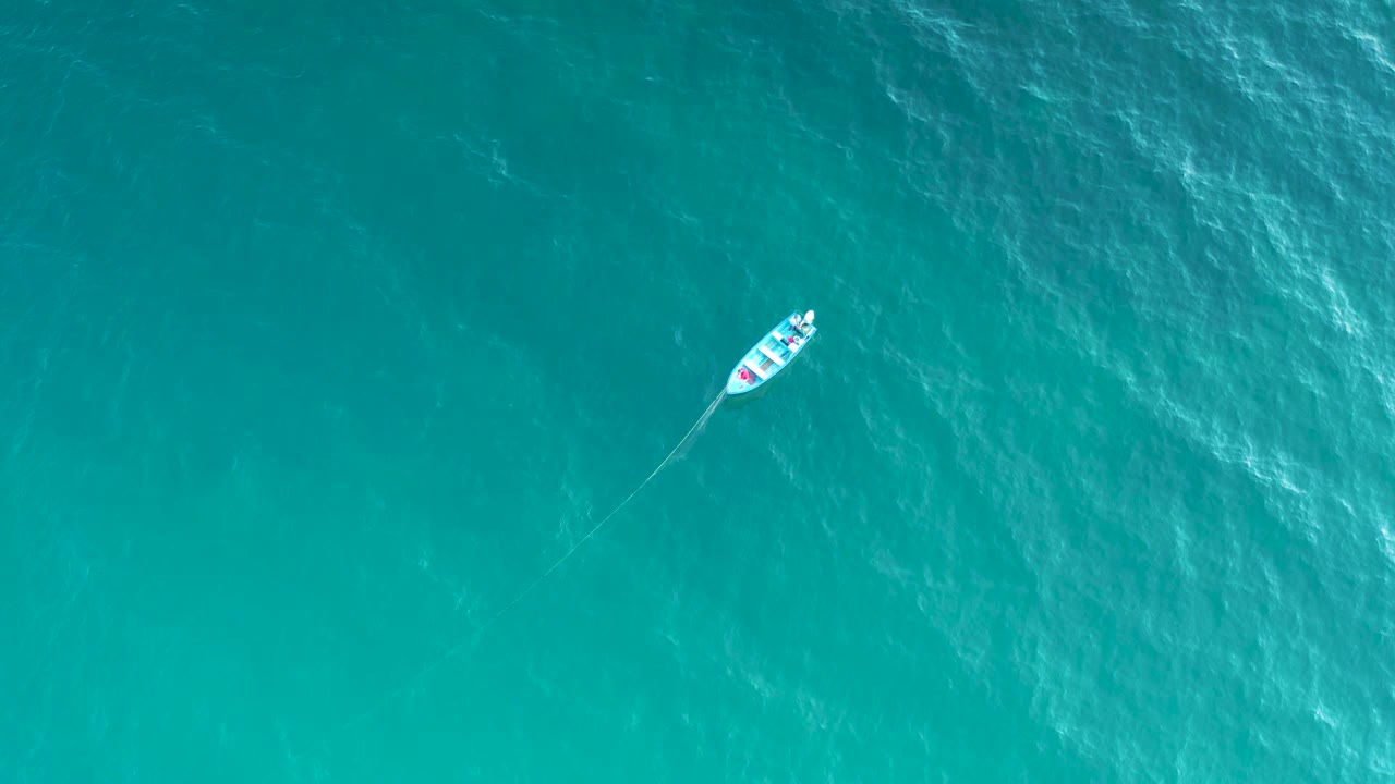 Overhead frame of the small boat slightly farther across open turquoise water, with pale wave texture behind it and no visible people.
