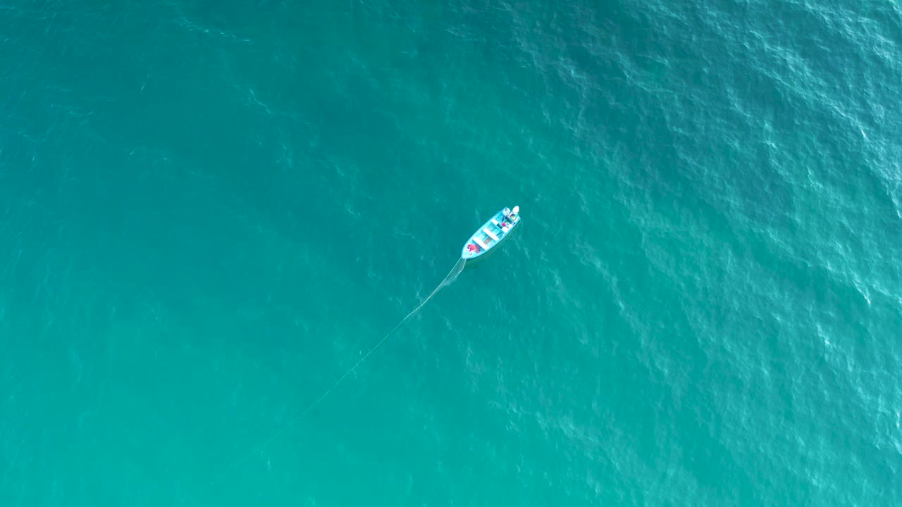 Overhead shot of a small boat left of center on blue green water, with a faint trailing line extending behind it and no visible people.