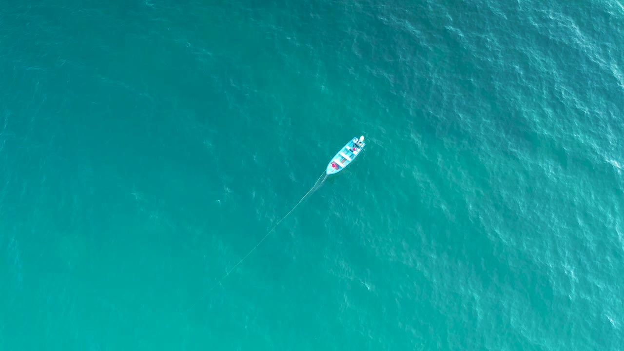 High overhead view of the white vessel slightly farther across blue-green sea in El Sargento La Ventana, Mexico, with a faint wake trailing behind and no visible people.