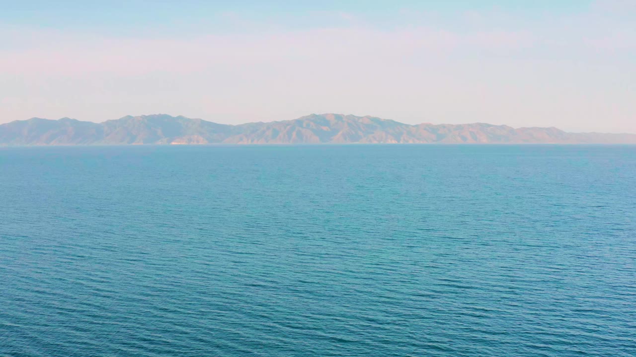 Opening frame of blue rippled water facing distant mountains in El Sargento La Ventana, Mexico, with a pale bright sky and no visible people.
