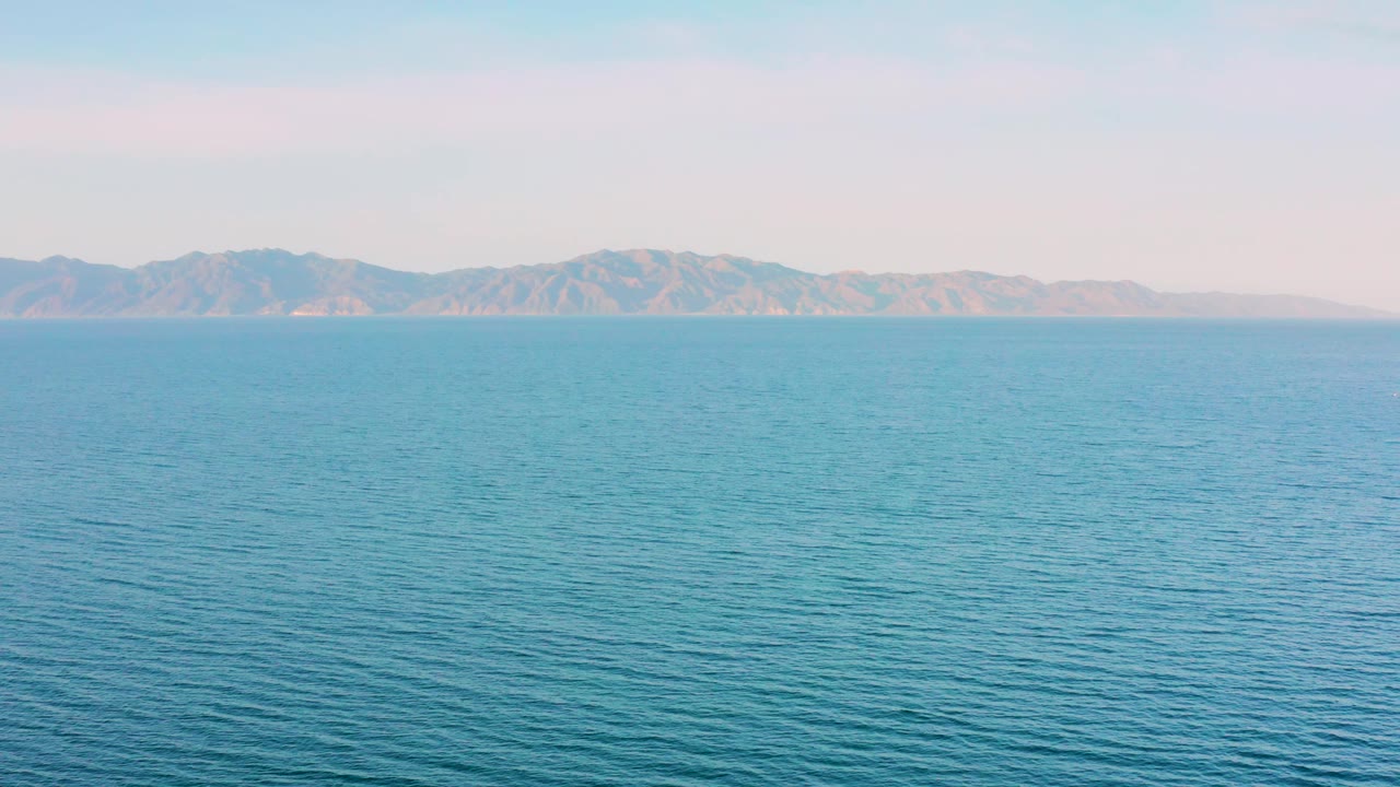 At 0:03, the sea remains evenly framed beneath a soft sky, with the mountain ridge stretching across the background and no visible people.
