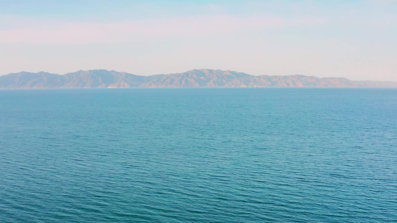A wide 4K frame from El Sargento La Ventana, Mexico with blue sea in the foreground, faint ripples across the water, and far mountains under soft light, no visible people.