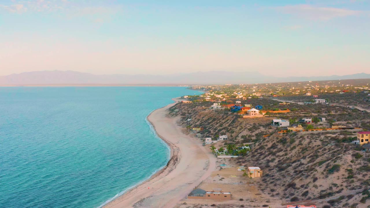 Aerial view in El Sargento La Ventana, Mexico showing turquoise water filling the left side, a narrow sand beach curving through the center, hillside homes on the right, and no visible people.