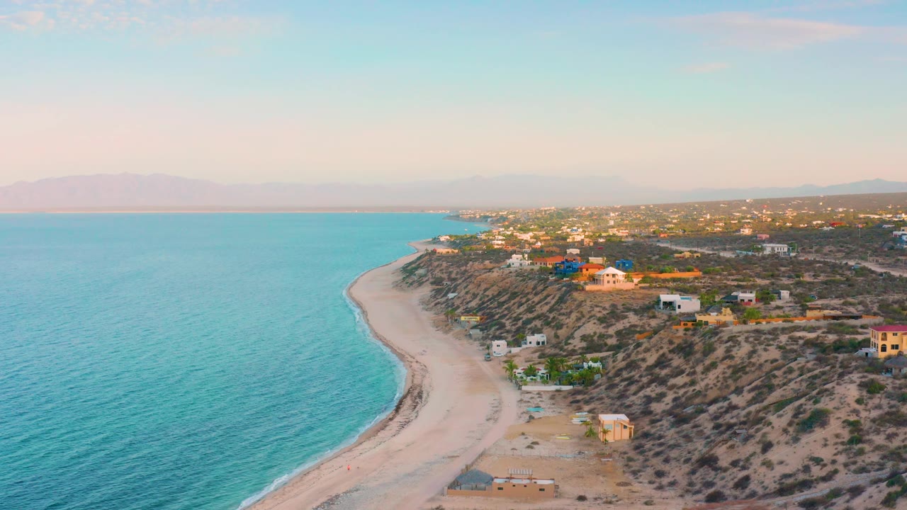 Wide aerial frame from El Sargento La Ventana, Mexico with calm blue sea at left, the shoreline bending below, hillside houses on dry terrain, distant mountains, and no visible people.