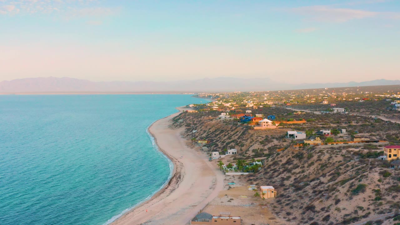 High coastal view with blue water, arcing beach, scrub hillside, small buildings, and soft sky.