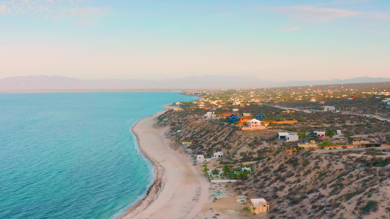 Opening aerial view over El Sargento La Ventana, Mexico, showing turquoise ocean on the left, a curving pale beach, dry coastal terrain, scattered homes, distant hills, and no visible people.