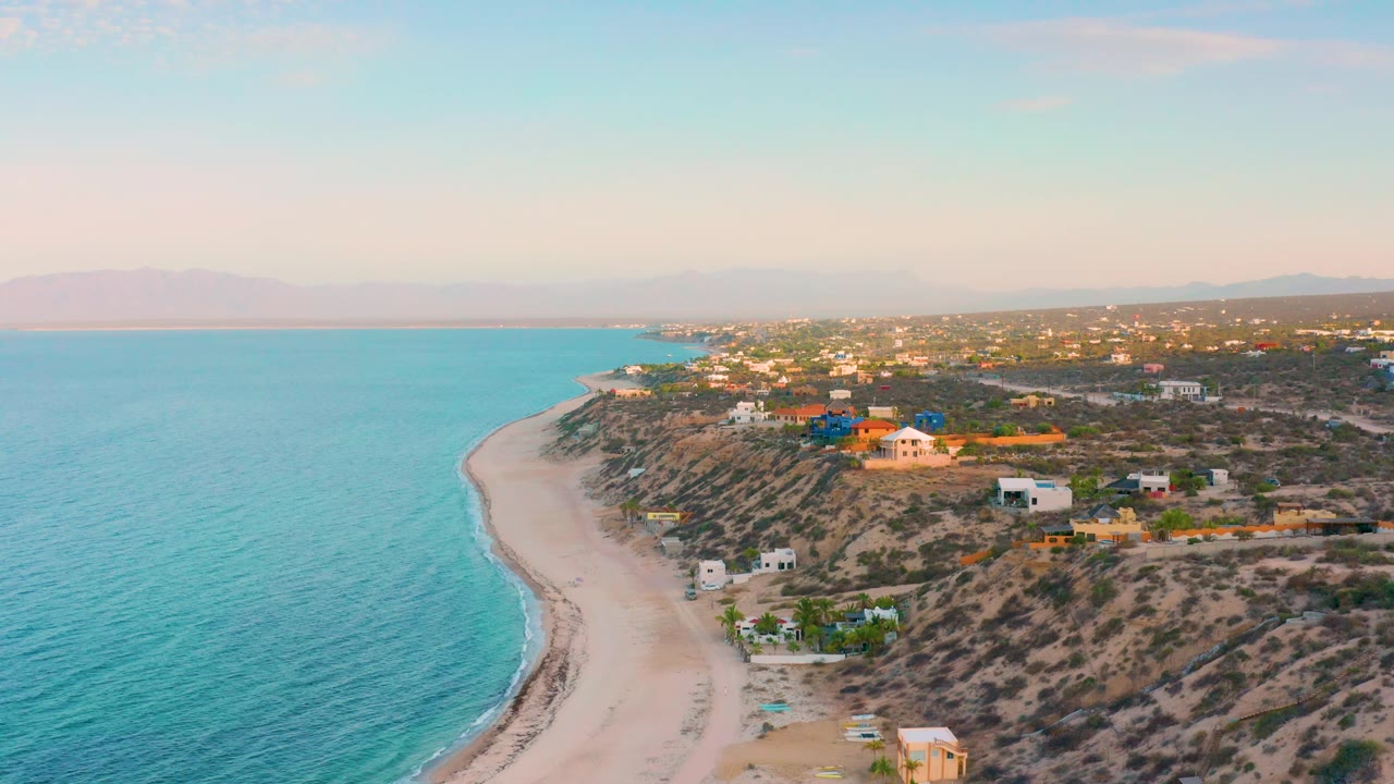 Drone-height view over El Sargento La Ventana with more open turquoise water filling the left frame, pale sand tracing the shore, low houses on the dry slope, and no visible people.