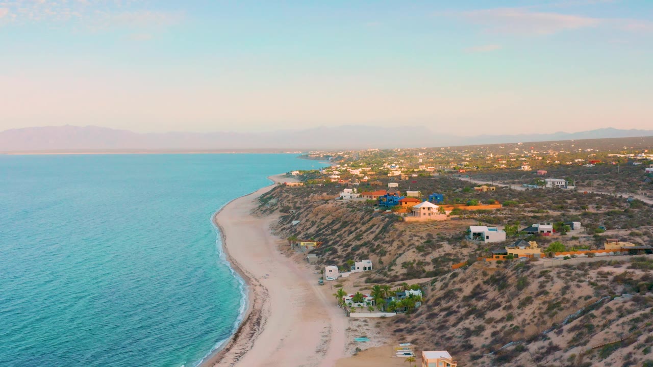 Coastal aerial with calm blue water, pale beach, and homes above the shore.