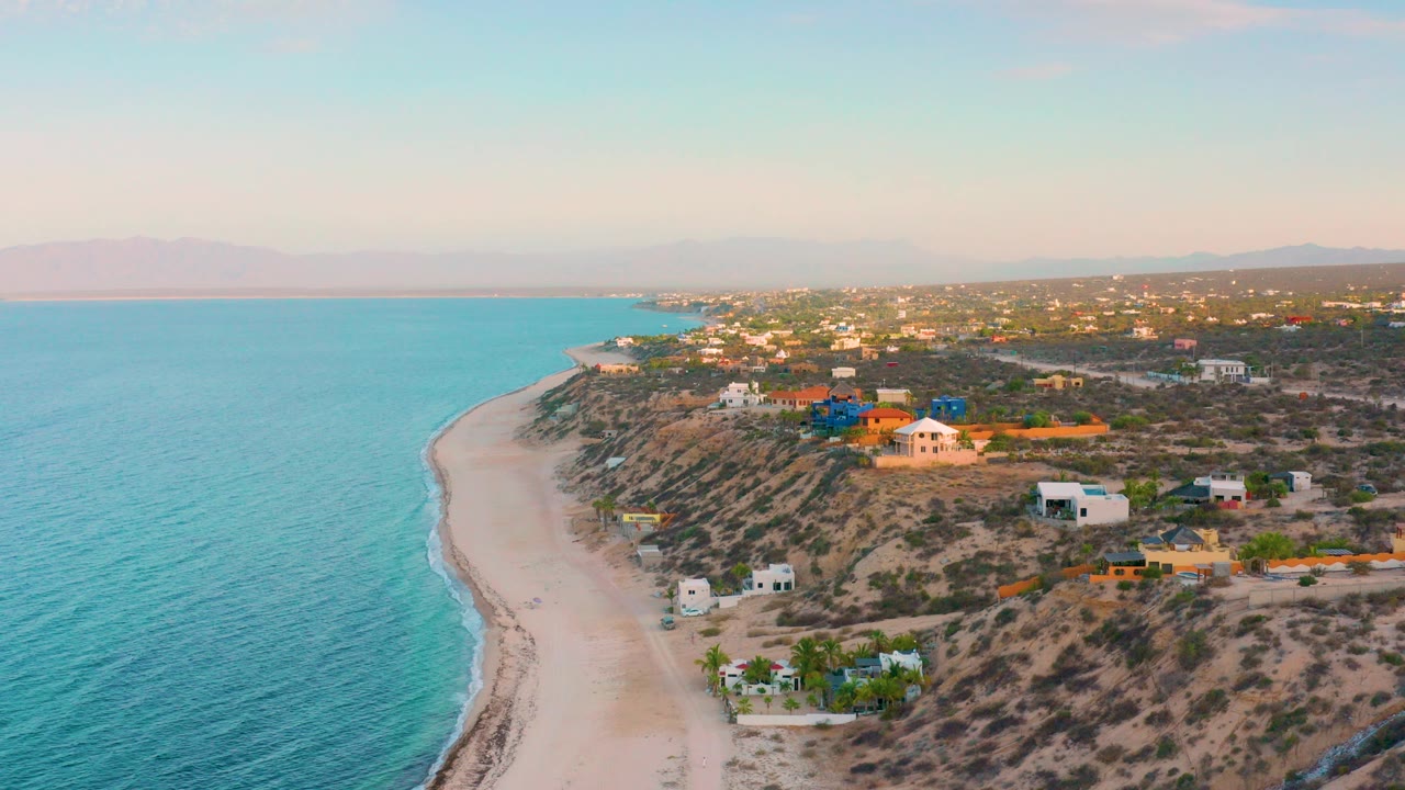 Drone view over El Sargento La Ventana, Mexico with turquoise water on the left, pale beach curving below, scattered hillside homes, distant mountains, and no visible people.