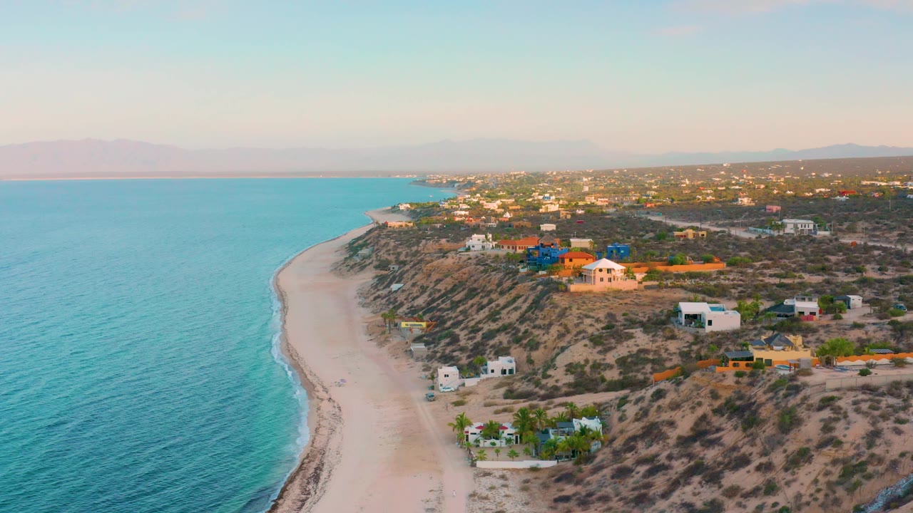 High drone view in El Sargento La Ventana, Mexico with more open ocean filling the left side, a pale beach below, hillside homes to the right, and no visible people.
