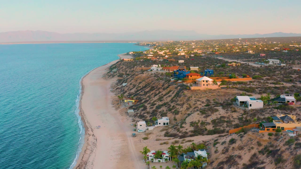 Drone view over El Sargento La Ventana, Mexico with the beach curve extending into the distance, clear blue water, hillside houses, and no visible people.