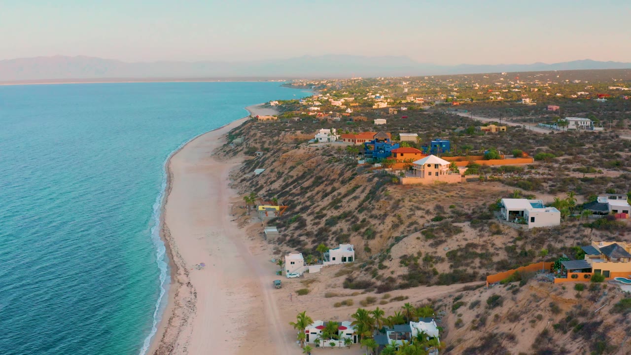 Wide aerial frame in El Sargento La Ventana, Mexico showing more open water at left, a narrow sandy shore, scattered homes on dry terrain, and no visible people.