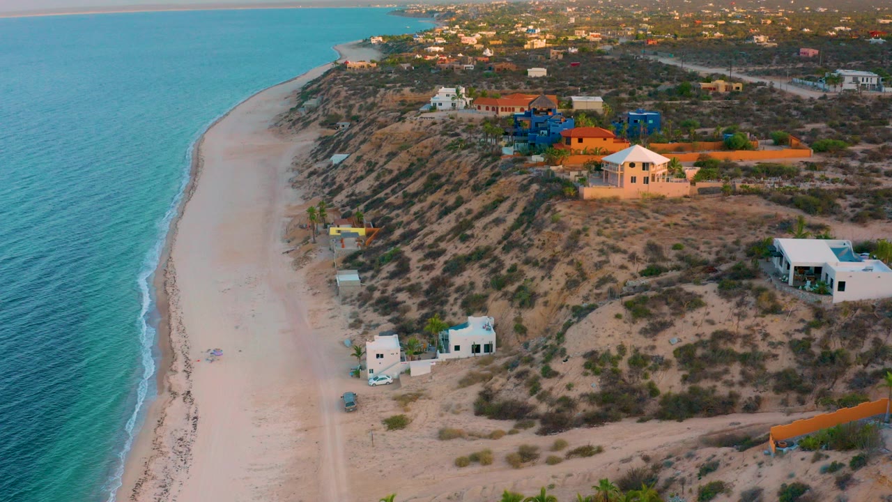 Aerial frame over El Sargento La Ventana, Mexico showing turquoise water beside a pale beach, scrub-covered dunes, modern coastal houses, and no visible people.