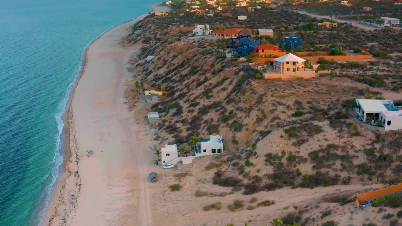El Sargento La Ventana, Mexico appears from above with turquoise sea, a narrow pale beach, dune vegetation, clustered coastal homes, warm light, and no visible people.