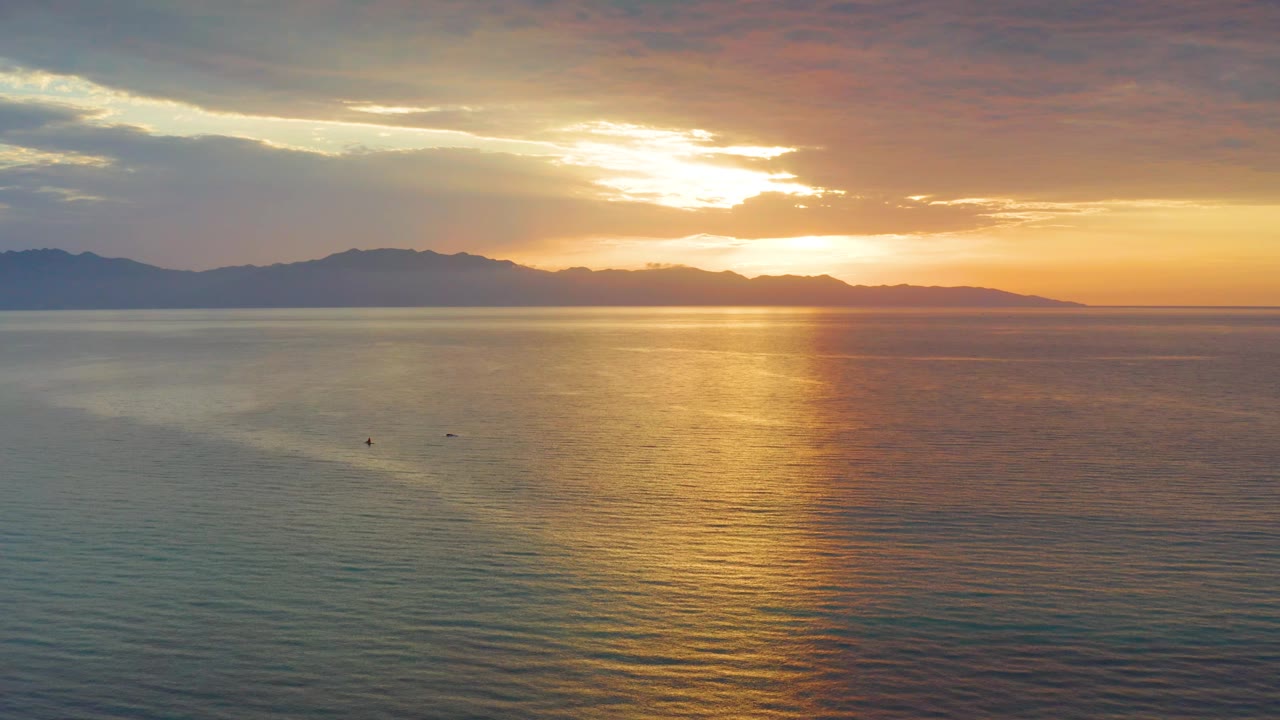 The sun sits behind a broken cloud layer over El Sargento La Ventana, Mexico, with orange sky, dark mountains, textured water, and no visible people.