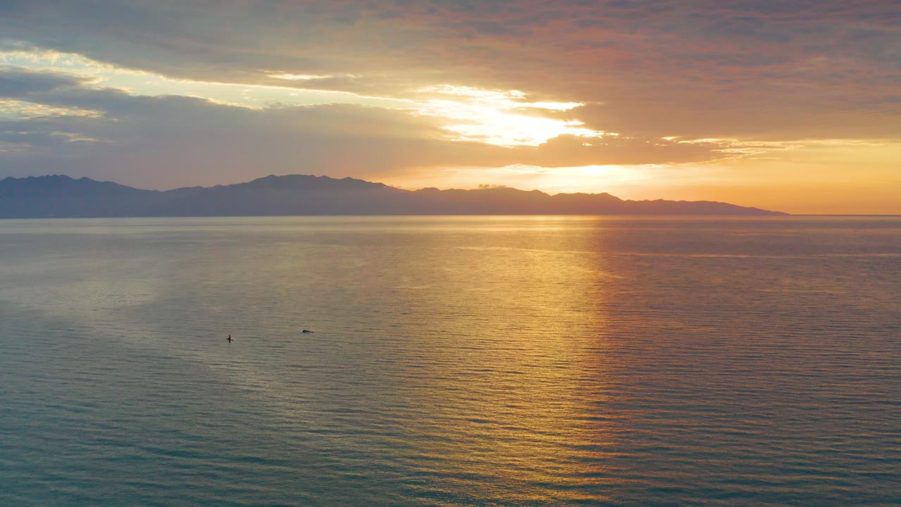 Low aerial sunset view over the ocean in El Sargento La Ventana, Mexico, with a broad gold reflection on the water, dark mountains on the horizon, and no visible people.