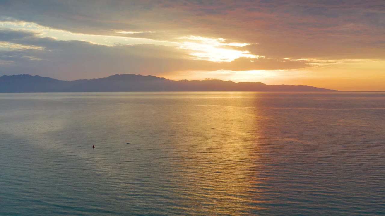 Wide low aerial frame over El Sargento La Ventana water, with the sun partly behind cloud, a vertical gold reflection, a dark mountain ridge, and no visible people.