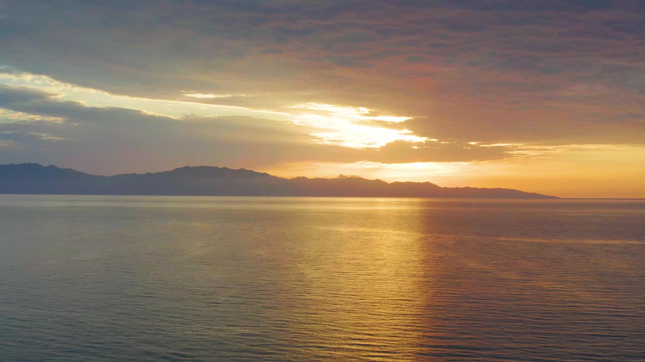 Second frame of the El Sargento La Ventana water view, with the sun partly hidden by clouds, a vertical gold reflection on the sea, distant land, and no visible people.