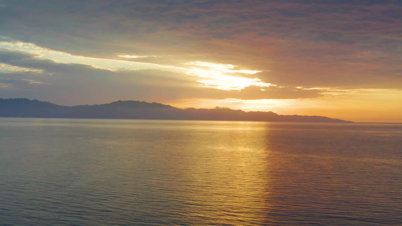 The sun sits behind broken clouds over El Sargento La Ventana, Mexico, with the reflection centered across calm water and no visible people.