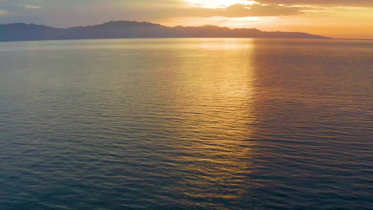 A broad dawn view shows the sun glow near the top of frame, a bright reflection on the water, and dark hills along the horizon in El Sargento La Ventana, Mexico, with no visible people.