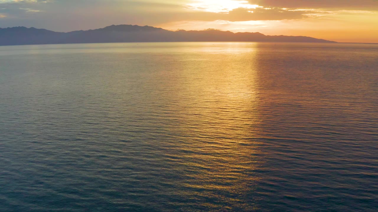 The water carries a vertical path of gold light toward low silhouetted hills under a warm dawn sky in El Sargento La Ventana, Mexico, with no visible people.