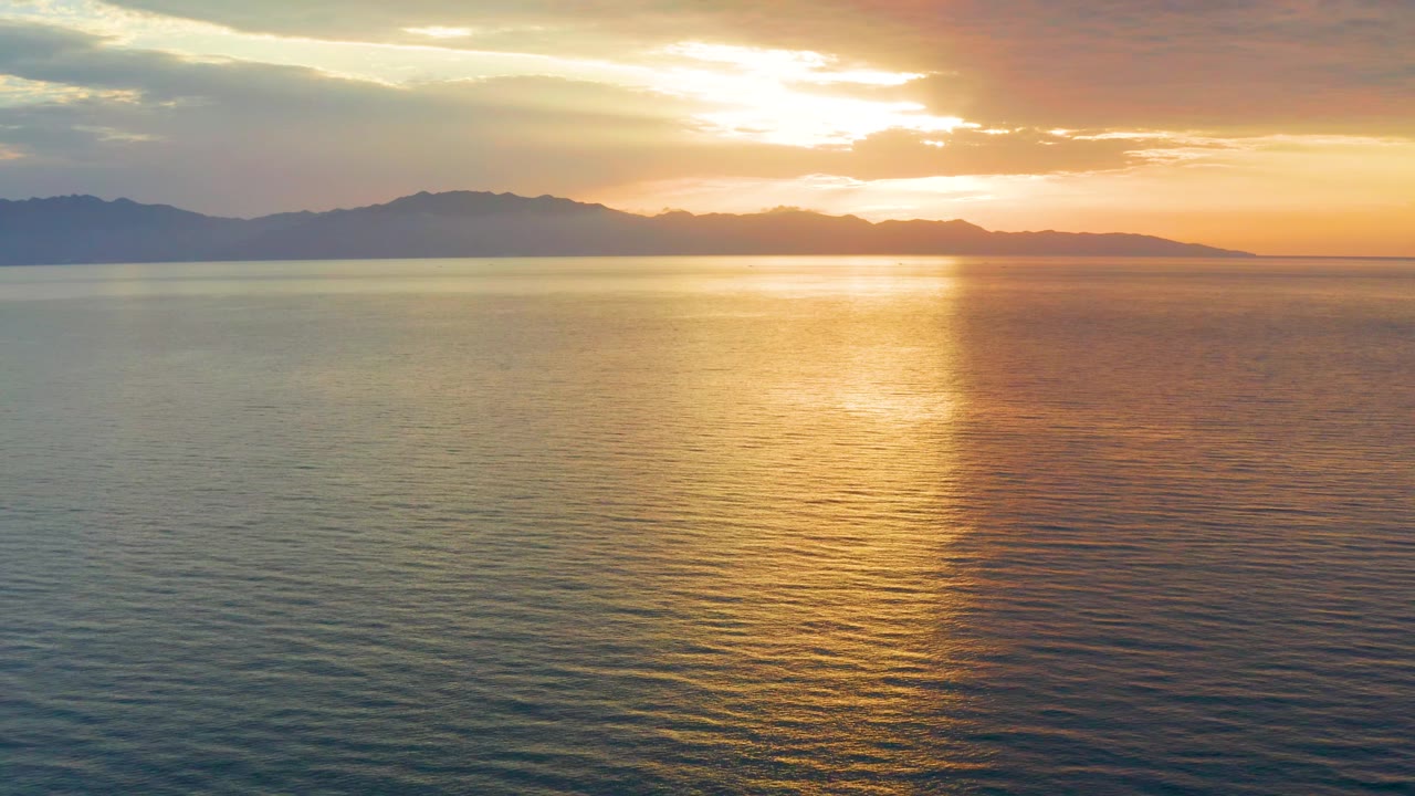 At 0:02, the mountain range stretches across the horizon while orange light spreads through cloud gaps and over the calm sea, with no visible people.