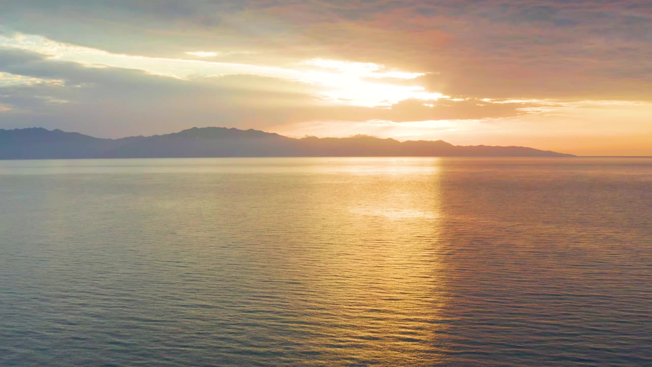 At 0:01, the sun sits behind bright cloud cover over El Sargento La Ventana, casting a wider gold reflection across lightly rippled water, with no visible people.