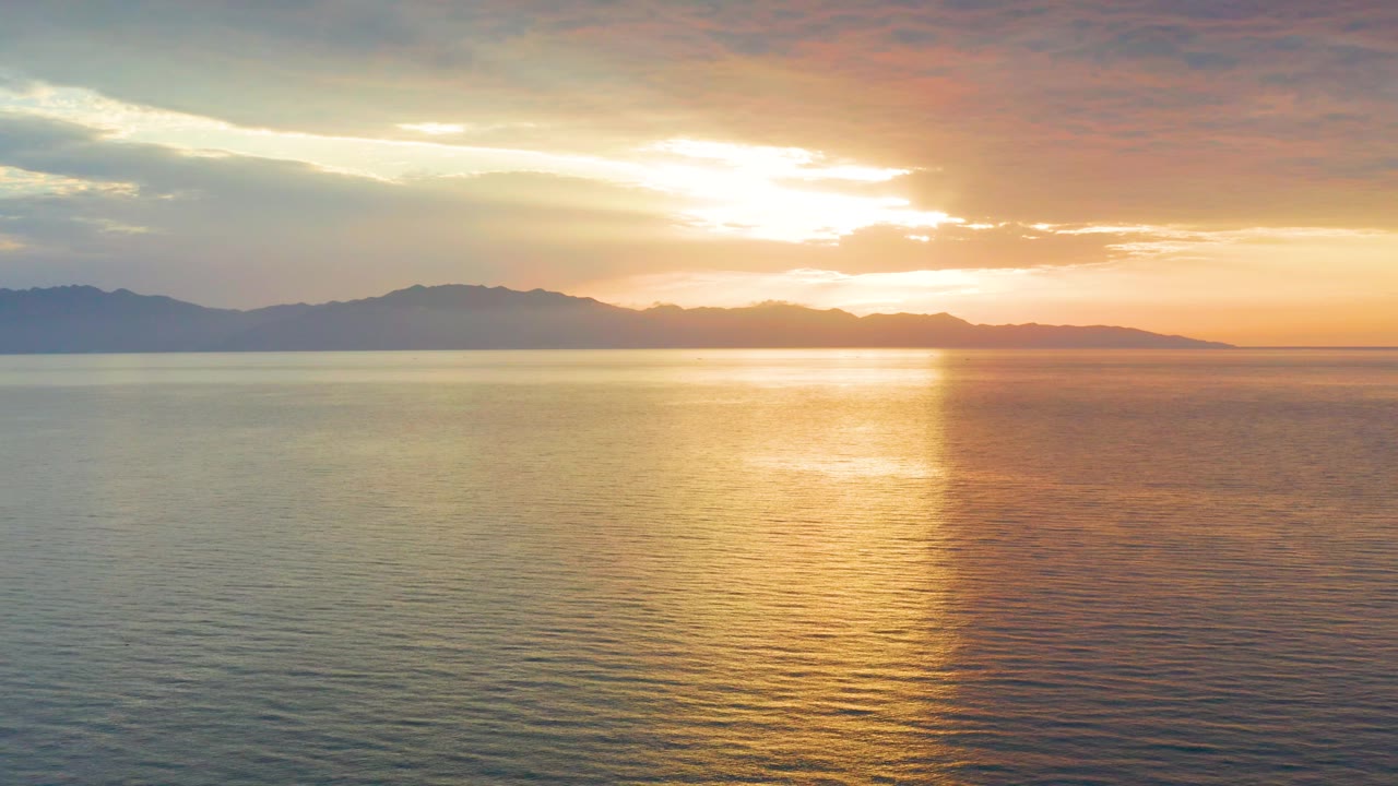 At 0:02, the low sun glows through orange cloud bands above El Sargento La Ventana, with a bright reflection column crossing the water and no visible people.