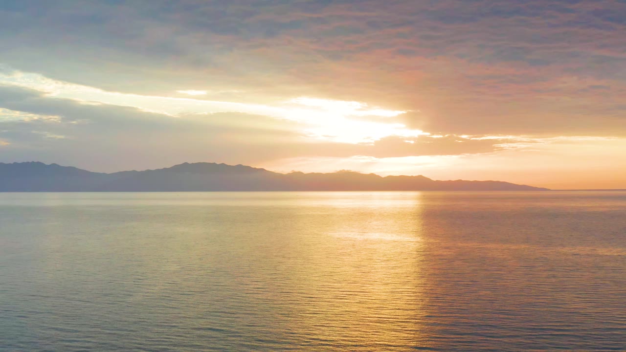 At 0:01, the sun sits just above the mountain line over El Sargento La Ventana, Mexico, spreading a vertical band of gold light across the calm sea with no visible people.