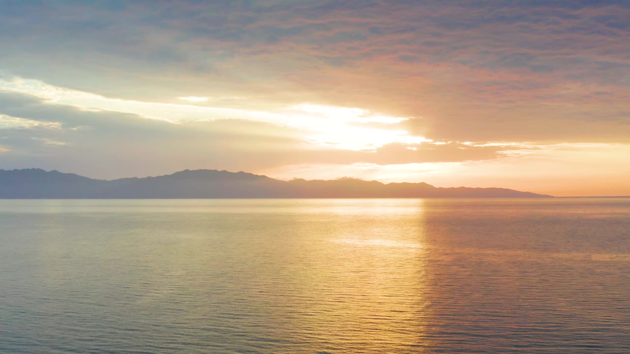 At 0:02, bright cloud-filtered sunlight hangs over the sea in El Sargento La Ventana, Mexico, with dark mountain silhouettes stretching across the background and no visible people.