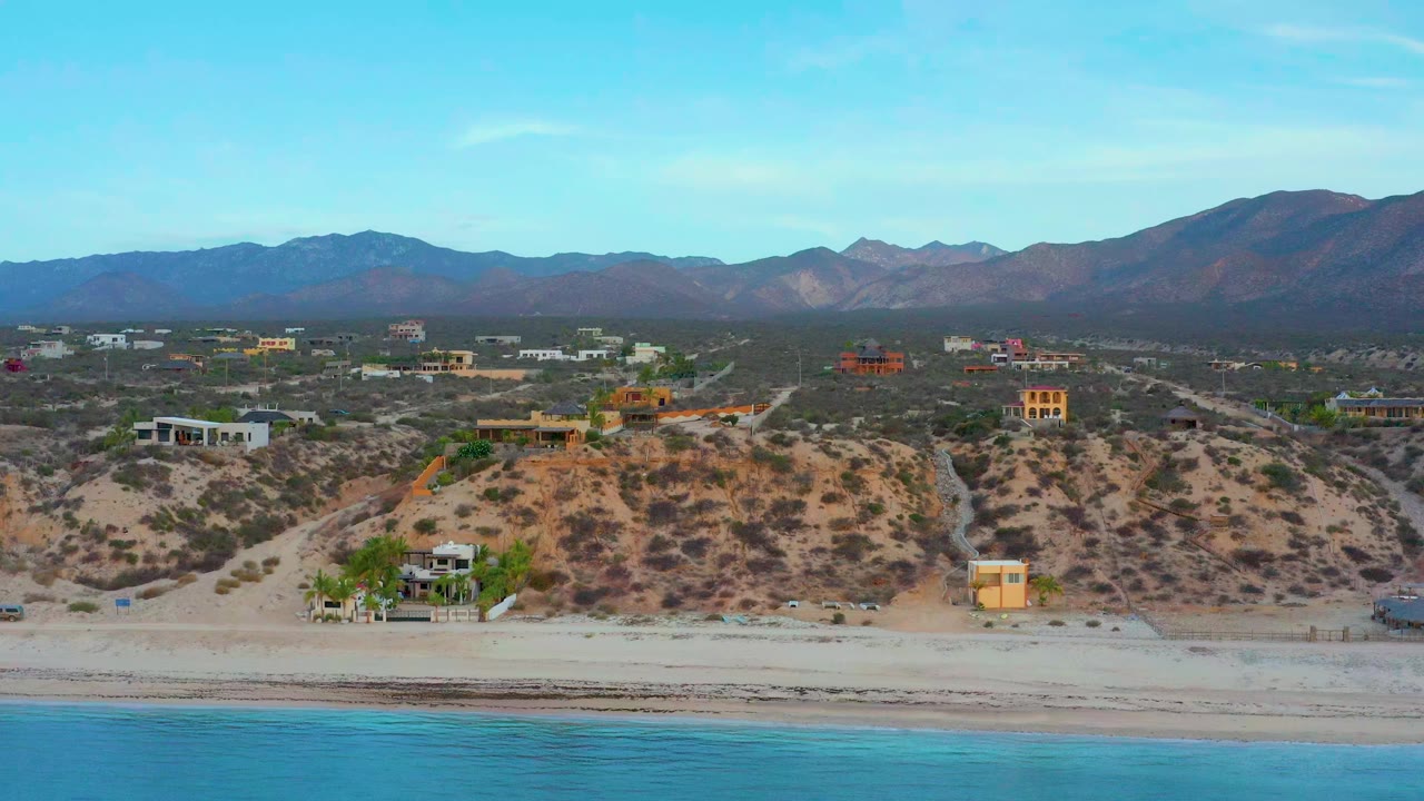 Wide aerial frame of El Sargento La Ventana, Mexico, with blue water at the bottom, a sandy beach, dry hillside lots, scattered homes, distant mountains, and no clearly visible people.