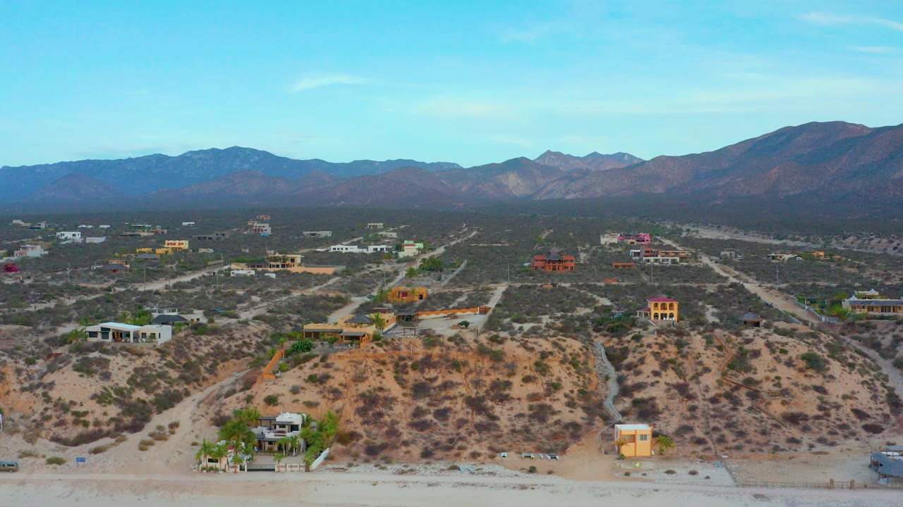 Wide aerial frame over El Sargento La Ventana, Mexico, with sandy foreground bluffs, scattered homes, dirt roads, distant mountains, blue sky, and no visible people.