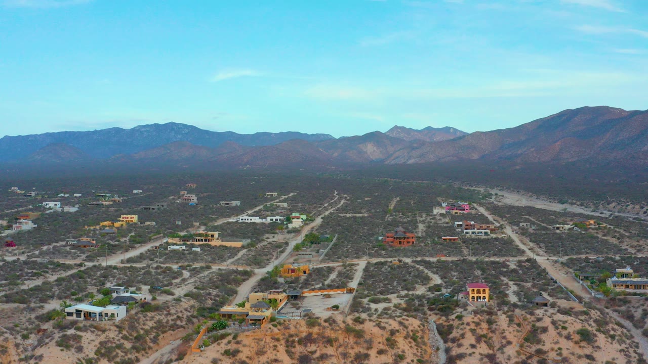 Opening aerial frame over El Sargento La Ventana, Mexico, showing desert scrub, pale dirt roads, colorful scattered houses, distant mountains, and no visible people.
