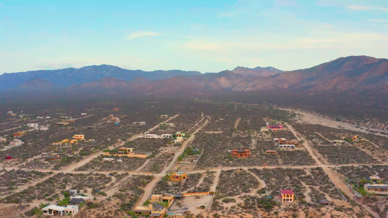 Opening aerial frame over El Sargento La Ventana, Mexico, showing pale sand streets, sparse desert vegetation, scattered homes, distant mountains, and no visible people.